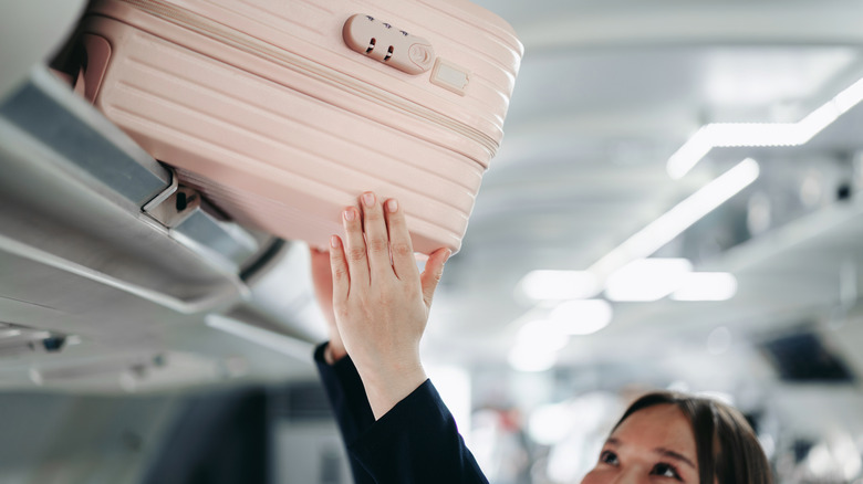 A woman stowing a suitcase in an overhead bin.